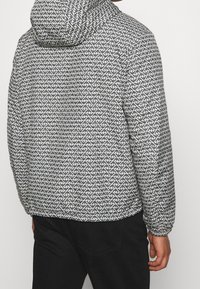 Vadderad jacka med huva, tillverkad av ett svart och vitt geometriskt mönstrat tyg. Elastiska muddar säkerställer en tät passform.