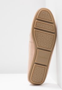 Semelle d'une chaussure Calvin Klein en daim beige reposant verticalement contre un mur blanc d'angle, montrant le logo en relief sur la semelle.