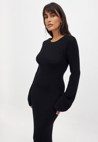 Robe noire en tricot côtelé avec de longues manches évasées et un col rond, présentant une silhouette ajustée qui s'étend jusqu'aux chevilles.