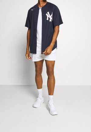 Hombre vestido con camiseta de béisbol de los New York Yankees en azul oscuro, camiseta blanca, pantalones cortos blancos, calcetines blancos y zapatillas blancas, de pie frente a un fondo liso.