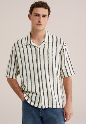 Mężczyzna w białej koszuli z krótkim rękawem z pionowymi czarnymi paskami i niebieskich dżinsach, stoi z jedną ręką w kieszeni na jednolitym tle.