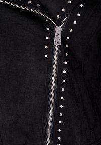 Veste en suède noir avec une fermeture à glissière argentée et des clous en métal le long d'un côté. Texture lisse avec une coupe ajustée.