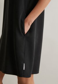 Black dress featuring a simple design, made from smooth fabric, with a side pocket and a small label at the hem.