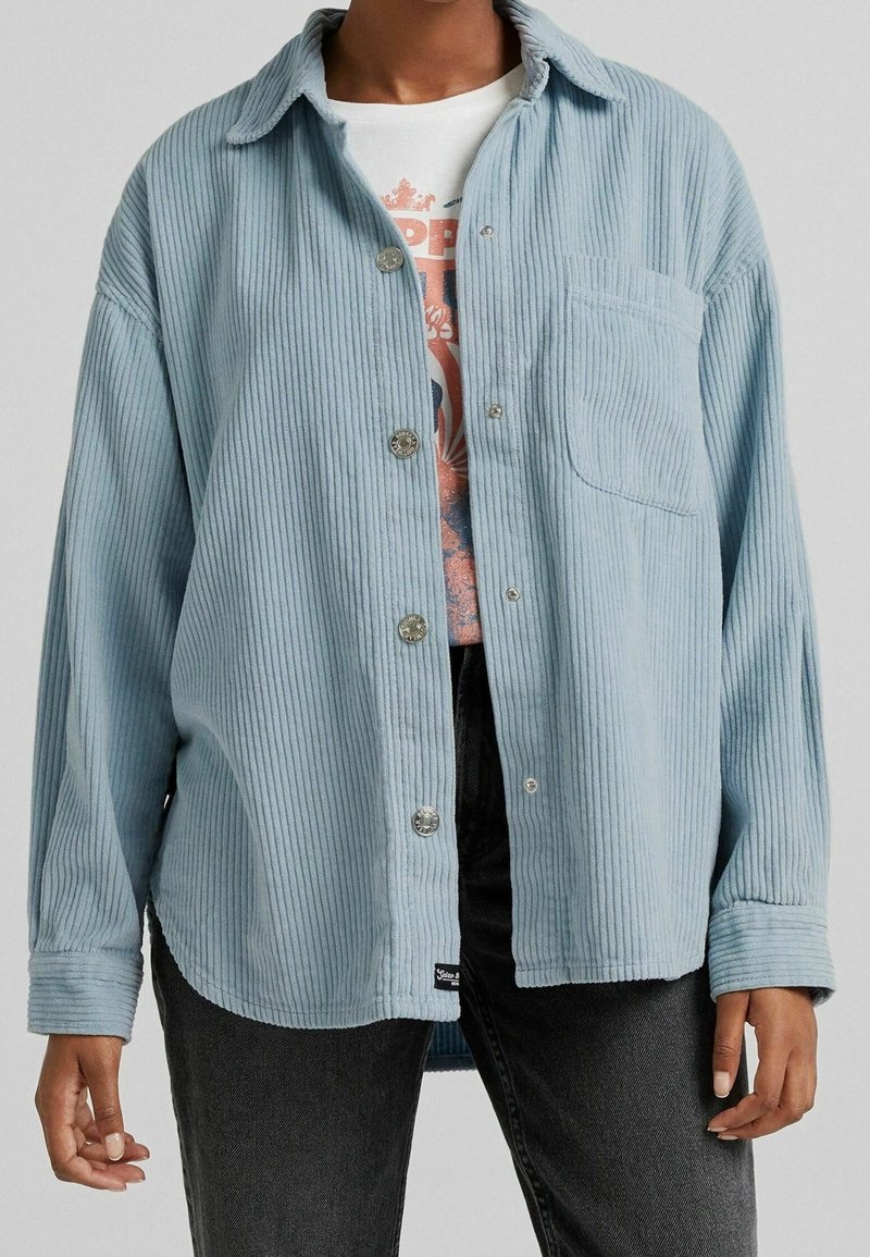 Personne portant une chemise bleu clair côtelée à boutons par-dessus un t-shirt blanc avec un motif graphique et un jean noir sur un fond uni.