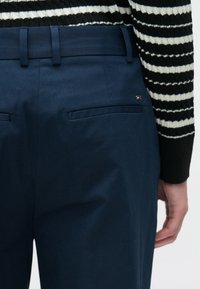 Μπλε παντελόνι με λείο ύφασμα, διαθέσιμο με δύο πίσω τσέπες και μικρή λεπτομέρεια με λογότυπο. Η μέση διαθέτει θηλιές για ζώνη για επιπλέον στήριξη.