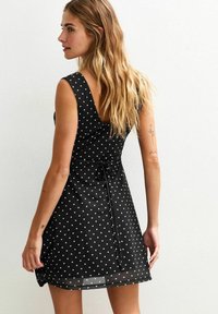 Robe sans manches noire avec des pois blancs. Présente un col en V et un lien à la taille. Texture fluide avec un ourlet légèrement transparent.