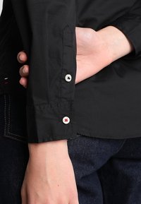 Černá košile s dlouhým rukávem s přiléhavým střihem, bílými knoflíky s červenými akcenty a hladkou texturou, nošená s tmavými džínami.