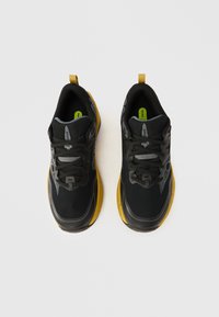 Ténis desportivos pretos com parte superior em rede, detalhes cinzentos, sola amarela e uma presilha para facilitar o calçar. Apresentam atacadores redondos e palmilhas almofadadas.