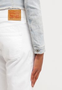 Persona che indossa jeans Levi's 501 bianchi e una giacca di jeans azzurro chiaro, con la mano appoggiata sul coscia su uno sfondo bianco.