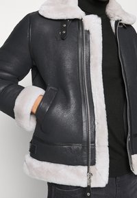 Grijze leren jas met witte schapenvachtvoering, ritsluiting en twee zijzakken. Bevat zwarte leren accenten en zilveren hardware.