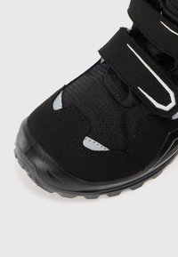 Zwarte wandelschoen met klittenbandsluitingen, bovenwerk van gestructureerde stof, rubberen zool en reflecterende accenten. Afgeronde teenvorm voor comfort en grip.