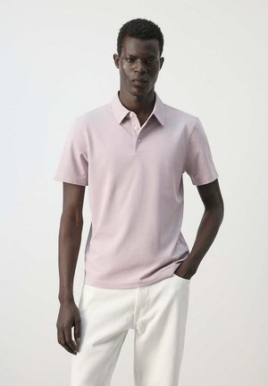 Hombre que lleva una camisa polo de manga corta de color rosa claro con pantalones blancos, de pie con una mano en el bolsillo frente a un fondo liso.