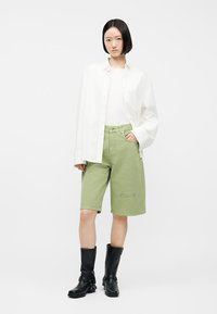 Naine, kes kannab valget nööpidega särki valge topi peal, helerohelisi põlveni ulatuvaid lühikesi pükse ja musti keskkõrge säärega saapaid, seisab ühe käega taskus.