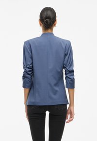 Skreddersydd blå blazer med en strukturert design, lange ermer og teksturert stoff. Har en pent krage og enkel bakseam. Stylet med svarte bukser.