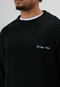 Zwarte fleece trui met een gestructureerde afwerking, ronde hals en een geborduurd "Young Athlete" logo in wit op de borst.