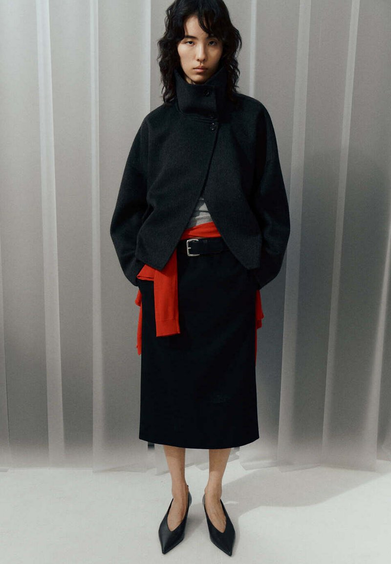 Femme portant une veste noire à col montant, une jupe midi noire, un haut rouge noué à la taille et des chaussures noires pointues, debout devant un rideau clair.