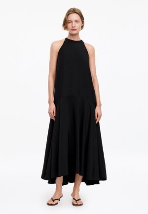 Frau steht und trägt ein ärmelloses schwarzes Neckholder-Kleid mit ausgestelltem asymmetrischem Saum und schwarze Sandalen vor weißem Hintergrund.