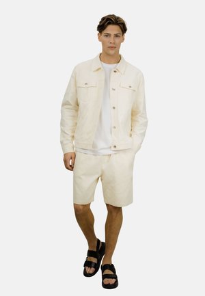 Ung mann står med hendene i lommene på en kremfarget jakke, iført matchende kremfargede shorts, hvit skjorte og svarte sandaler på hvit bakgrunn.