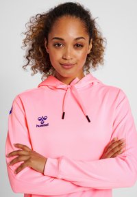 Spīdīgi rozā kapuce izgatavota no mīksta auduma, ar logotipu uz krūtīm un regulējamām auklām. Gluda tekstūra ar garām piedurknēm.