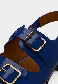 Sandale bleue brillante avec deux brides à boucles en métal doré et semelle intérieure marron, montrée en gros plan sur fond blanc.