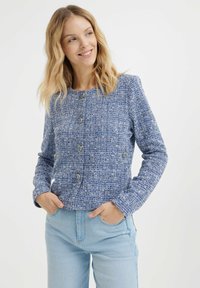 Giacca di tweed blu con una finitura testurizzata, caratterizzata da un collo tondo, maniche lunghe e bottoni color oro. Indossata sopra jeans blu chiaro.