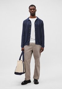 Kék kötött kardigán gombos elől, fehér pólóval és bézs nadrággal párosítva. Krém és kék vászontáska van a kezében. Fekete cipő.