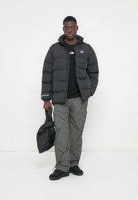Schwarze Pufferjacke mit Kapuze, Reißverschluss und Logodetail. Das Modell trägt olivfarbene Cargo-Hosen und hält eine kleine schwarze Jacke.