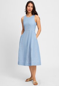 Lichtblauw mouwloos jurkje met een nauwsluitende corsage, uitlopende rok, zakken aan de zijkanten en een gestructureerde stof. Draagt bij flatbeige sandalen.