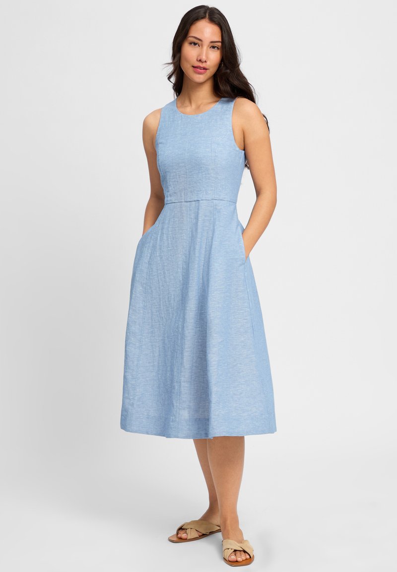 Lichtblauw mouwloos jurkje met een nauwsluitende corsage, uitlopende rok, zakken aan de zijkanten en een gestructureerde stof. Draagt bij flatbeige sandalen.