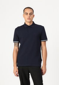 Polo majica v mornarsko modri barvi, izdelana iz teksturirane tkanine, z ovratnikom, tremi gumbi in manšetami z belimi črtami. Rdeč logotip na sprednji strani.