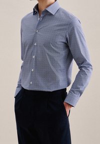 Muž oblečený v modré vzorované košili s dlouhým rukávem, zasunuté do tmavě modrých kalhot, s rukama v kapsách, na béžovém pozadí.