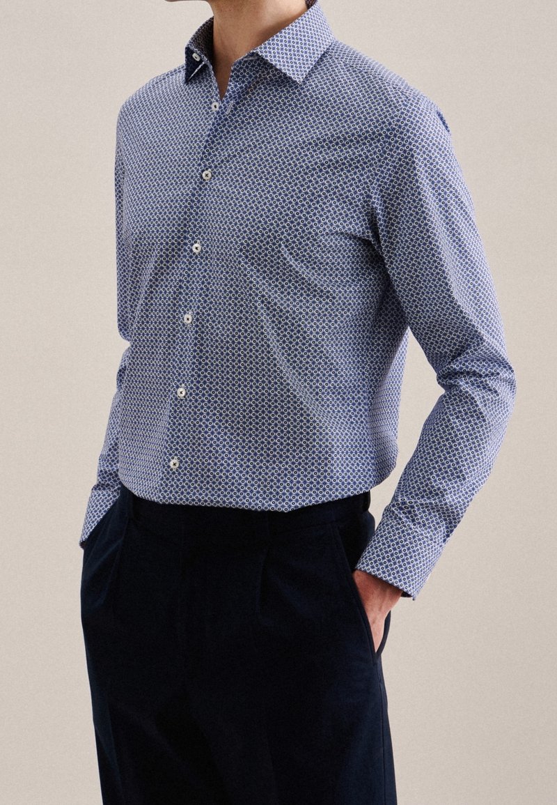 Muž oblečený v modré vzorované košili s dlouhým rukávem, zasunuté do tmavě modrých kalhot, s rukama v kapsách, na béžovém pozadí.