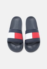 Σαγιονάρες σε ναυτικό μπλε με δύο οριζόντιες λουρίδες: μία κόκκινη και μία λευκή με κόκκινη λεπτομέρεια, κατασκευασμένες από υφή ελαστικού. Λογότυπο στη σόλα.