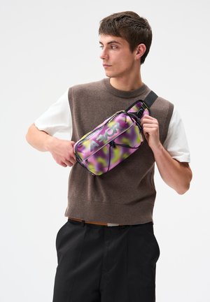 Jeune homme ajustant un sac bandoulière AEOR violet et jaune, portant un pull sans manches marron par-dessus une chemise blanche et un pantalon noir sur fond blanc.