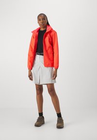 Veste imperméable rouge avec capuche, poignets élastiques et logo ; t-shirt noir en dessous, shorts gris clair, chaussures de randonnée marron.