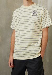 Gulrandig T-shirt med grå ränder, med en cirkulär Nike Athletic Club-logotyp på bröstet, tillverkad av mjukt tyg. Avslappnad passform.
