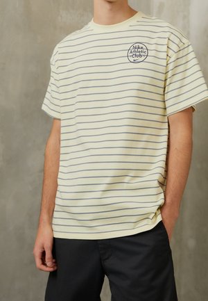 Gulrandig T-shirt med grå ränder, med en cirkulär Nike Athletic Club-logotyp på bröstet, tillverkad av mjukt tyg. Avslappnad passform.