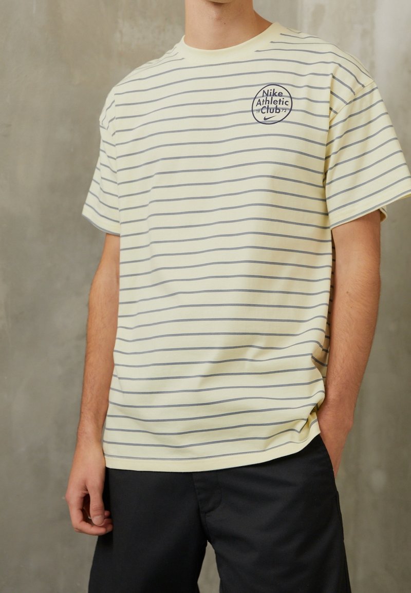 Gulrandig T-shirt med grå ränder, med en cirkulär Nike Athletic Club-logotyp på bröstet, tillverkad av mjukt tyg. Avslappnad passform.