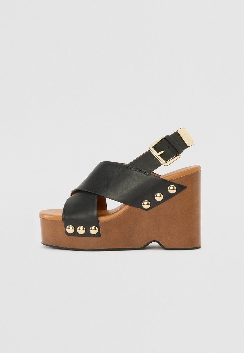 Sandales compensées en cuir noir avec un design à lanières croisées, des clous métalliques et une semelle plateforme beige. Boucle ajustable sur la lanière de la cheville.