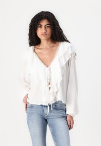 Blusa bianca con bordi arricciati, scollo a V profondo e dettagli in pizzo al colletto. Maniche lunghe con vestibilità rilassata. Abbinata a jeans azzurri.