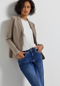 Beige kavaj med figursydd passform, vit skjorta och blå jeans. Kavajen har två knappar och fickor; jeansen har en klassisk passform och sömdekor.
