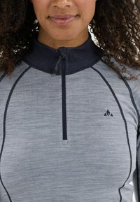 Harmaa vetoketjullinen paita, jossa on tumma kaulus ja saumat. Vasemmalla puolella on erottuva logo. Materiaali näyttää tekstuuriltaan kevyeltä.