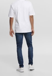 Bílý tričko s krátkým rukávem, volně střižené, v kombinaci s tmavě modrými džínami a bílými teniskami. Hladká bavlněná látka, jednoduchý design.