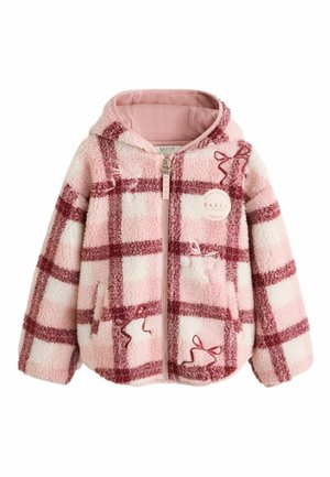 Pige med krøllet hår iført en pink og rød ternet lynlåsjakke, brede blå jeans og hvide sneakers, der står på et gråt gulv.