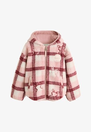 Pige med krøllet hår iført en pink og rød ternet lynlåsjakke, brede blå jeans og hvide sneakers, der står på et gråt gulv.