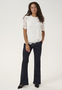 White lace top with short sleeves, featuring a scalloped hem, paired with dark blue flared jeans and black pointed-toe heels.