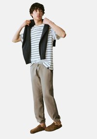 Camiseta de rayas blanca y negra, pantalones de chándal grises, suéter negro colgado sobre los hombros y zuecos marrones. Atuendo casual y cómodo.