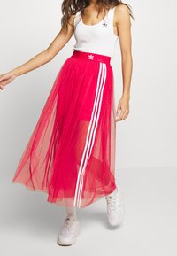 Red tulle skirt with white stripes down the sides, paired with a white tank top featuring a black logo. The skirt is ankle-length and sheer.