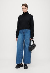 Pulover negru cu guler înalt și textură cordată, asociat cu blugi largi, de culoare albastră. Accesoriile includ o mică geantă neagră cu ținte și pantofi negri, masivi.