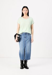 Hellgrünes T-Shirt mit kurzen, umgeschlagenen Ärmeln, kombiniert mit einem blauen Jeans-Maxikleid mit einem Schlitz vorne. Schwarze ankle boots und eine kleine schwarze Tasche.